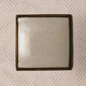 May include: Square ceramic coaster with a light gray center and a dark brown border. The coaster has a glossy finish and is likely handmade. The square shape is simple and modern, suitable for various home decor styles.