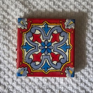May include: A red, white, blue, and yellow ceramic tile with a geometric pattern. The tile has a floral design with a central flower and four surrounding leaves.