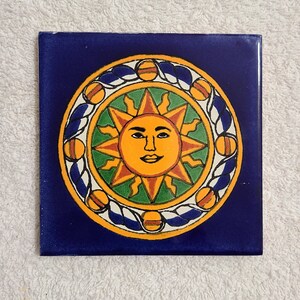 May include: A blue ceramic tile with a yellow sun with a face in the center. The sun is surrounded by a green, orange, and white design.