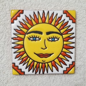 May include: A yellow ceramic tile with a hand-painted sun face. The sun has a red and orange halo with black outlines. The sun has blue eyes, black eyebrows, and a red mouth.
