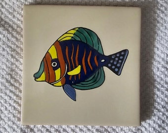 May include: A ceramic tile with a hand-painted design of a colorful fish with yellow, orange, blue, and green stripes. The fish is swimming to the right.