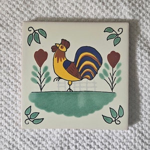 May include: A white ceramic tile with a hand-painted rooster design. The rooster is yellow, brown, blue, and red. The tile has a green background and a white border.