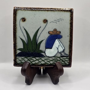 May include: A hand-painted ceramic tile with a blue and white illustration of a person wearing a hat sitting in front of a green plant. The tile is on a wooden stand.