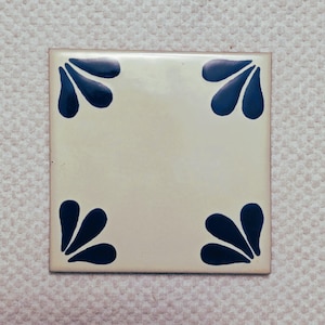 May include: Square ceramic tile with a cream-colored background and navy blue leaf-like designs in each corner. The tile has a glossy finish and is likely for decorative use.