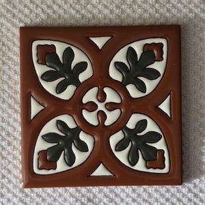 May include: A square ceramic tile with a brown background and a white and green geometric design. The design features four stylized leaves with a central circle.