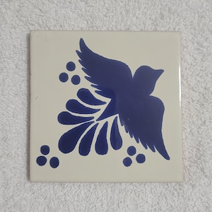 May include: A white ceramic tile with a blue bird design. The bird has a long tail with a floral pattern. The tile is perfect for adding a touch of Mexican style to your home.