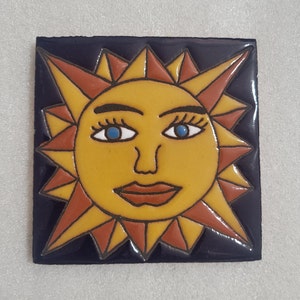 May include: A ceramic tile with a stylized sun face. The sun has a yellow face with a brown outline, blue eyes, and a brown mouth. The sun's rays are red and yellow.