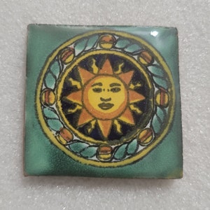 May include: A green ceramic tile with a yellow sun with a face in the center. The sun is surrounded by a yellow and green wreath with orange and red accents.