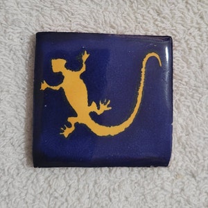 May include: A blue ceramic tile with a yellow silhouette of a lizard with a long tail.