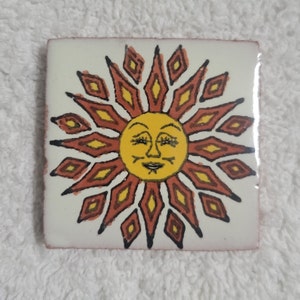 May include: Square ceramic tile featuring a smiling sun design. The sun has a yellow face with closed eyes and a smiling mouth. The sun's rays are a combination of orange, yellow, and black diamond shapes, set against a white background.