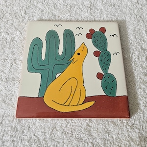 May include: A yellow coyote with a green cactus and prickly pear cactus on a white tile. The tile has a brown border.
