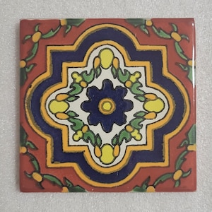 May include: A square ceramic tile with a colorful geometric pattern. The tile features a central blue flower with yellow accents, surrounded by a red, yellow, and blue design.