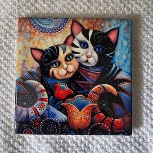 May include: A ceramic tile featuring a colorful illustration of two cats with intricate patterns and a stylized flower. The cats are black, white, and blue, and the flower is orange, red, and blue.