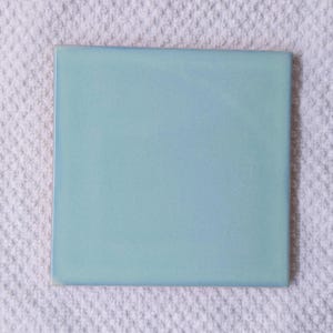 May include: A square, light blue ceramic tile with a glossy finish. The tile has a slightly darker blue edge and is set against a white, textured background. This tile is suitable for home decor projects.