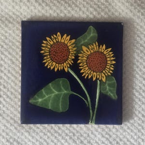 May include: A blue ceramic tile with two yellow sunflowers with brown centers and green leaves on a dark blue background.