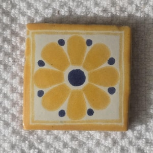 May include: A yellow ceramic tile with a blue and white floral design. The tile has a square shape and a white background.