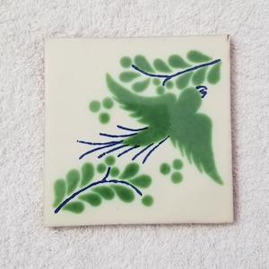 May include: A white ceramic tile with a green bird design. The bird is flying with its wings spread and has a long tail. The tile is decorated with green leaves and blue outlines.