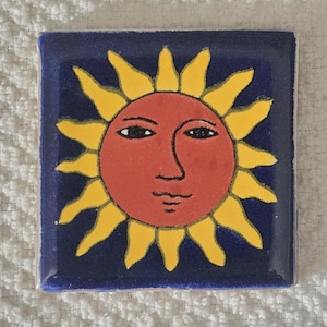 May include: A ceramic tile with a yellow sun with a red face and black eyes on a blue background.