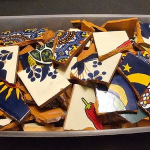 May include: A clear plastic bin filled with broken ceramic tiles. The tiles feature various designs, including floral patterns, a sun, a star, and a chili pepper. The color palette includes blue, white, yellow, red, and green.