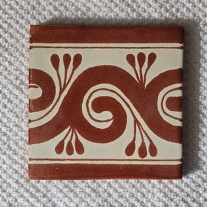 May include: A square ceramic tile with a white background and a brown design. The design features a repeating pattern of stylized swirls and leaves.