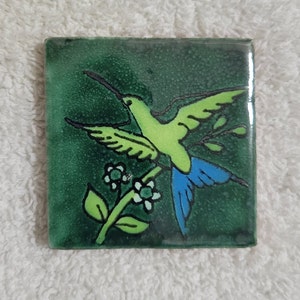 May include: Square ceramic tile with a hand-painted hummingbird design. The bird is lime green with blue tail feathers, perched on a branch with white flowers. The background is a dark green with a textured appearance.