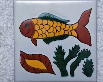 May include: Square ceramic tile featuring a hand-painted fish design. The fish has a yellow body with orange scales, a red head and tail, and green fins. Additional elements include a leaf and seaweed in shades of red, yellow, and green.