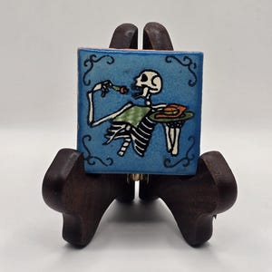 May include: A blue ceramic tile with a hand-painted design of a skeleton eating a meal. The skeleton is wearing a green and white striped shirt and holding a plate with food. The tile is on a wooden stand.