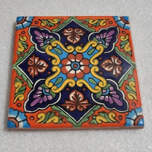 May include: A colorful ceramic tile with a geometric pattern of flowers and leaves in shades of blue, green, yellow, orange, red, and purple. The tile has a white background and a dark blue border.