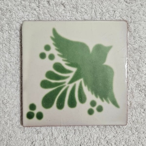 May include: A white ceramic tile with a green stylized bird design. The bird is in the corner of the tile and has a wing spread out. There are three green dots on each side of the bird.