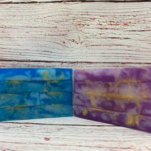 May include: Two bars of handmade soap with a marbled design. One bar is blue with white and gold swirls, and the other is purple with white and gold swirls.