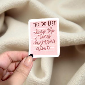 May include: A pink sticker with the words "TO DO LIST" at the top, followed by "-keep the tiny humans alive" in a handwritten style. The sticker is held by a hand with a French manicure against a cream-colored fabric background.