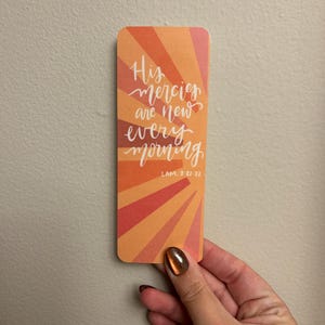 May include: A rectangular bookmark with a gradient orange and red sunburst design. The bookmark features the text "His mercies are new every morning" in white script, with the reference "LAM. 3:22-23" below. The bookmark is held by a hand.