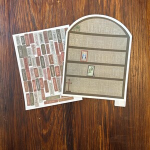 K&ouml;nnte beinhalten: Zwei Papierausstiche auf einer Holzoberfl&auml;che. Einer ist ein B&uuml;cherregal-Design mit einem Kreuz und gerahmter Kunst. Der andere ist eine Sammlung rechteckiger Etiketten mit Text. Die Etiketten tragen Namen wie "Genesis", "Exodus" und "Johannes".