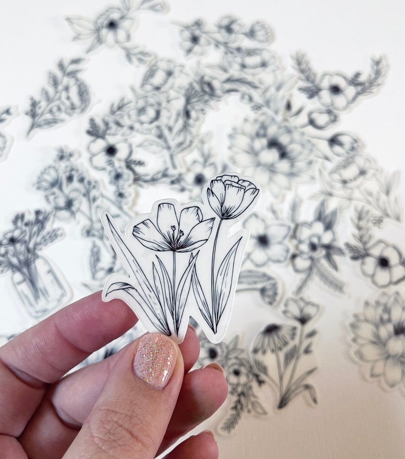 Puede incluir: Una hoja de pegatinas transparentes con dibujos de l&iacute;nea negra de varias flores y arreglos florales. Las pegatinas est&aacute;n dispuestas en un patr&oacute;n aleatorio sobre una superficie blanca.