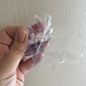 Puede incluir: Una pegatina transparente con un dise&ntilde;o floral en blanco y negro. El dise&ntilde;o presenta una flor grande con flores m&aacute;s peque&ntilde;as y hojas que la rodean. La pegatina est&aacute; sobre un fondo blanco.