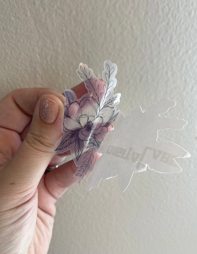 Puede incluir: Una pegatina transparente con un dise&ntilde;o floral en blanco y negro. El dise&ntilde;o presenta una flor grande con flores m&aacute;s peque&ntilde;as y hojas que la rodean. La pegatina est&aacute; sobre un fondo blanco.