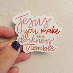 May include: A sticker with the words "Jesus you make the darkness tremble" in a cursive font. The text is in shades of pink, orange, and red. The sticker is held by a hand with a dark purple nail polish.