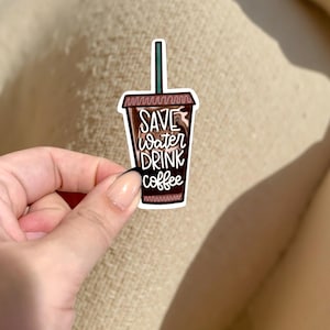 May include: A sticker featuring a coffee cup design with the text "SAVE water DRINK coffee" in white script. The cup is brown with a green straw and a white border. The sticker is held by a hand.