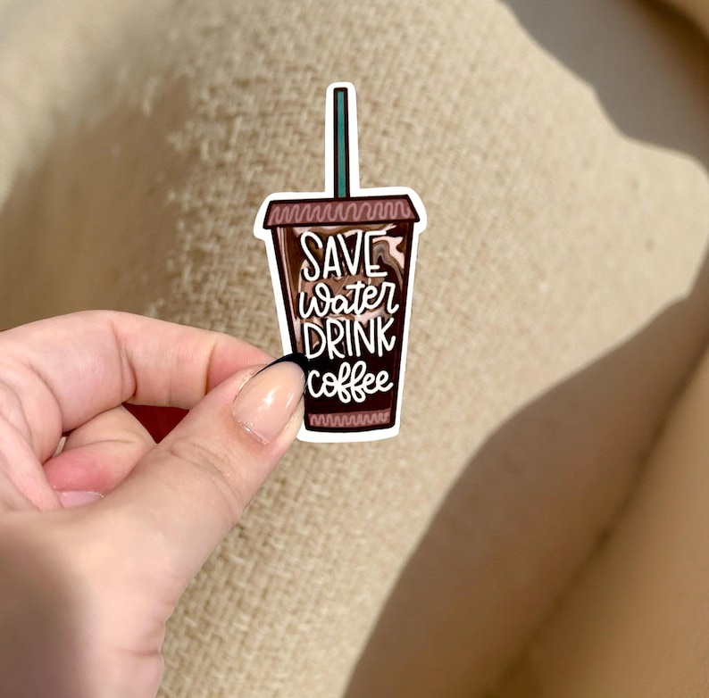 May include: A sticker featuring a coffee cup design with the text "SAVE water DRINK coffee" in white script. The cup is brown with a green straw and a white border. The sticker is held by a hand.