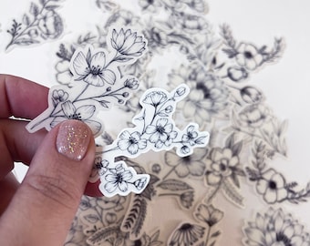 May include: A sheet of clear stickers with black line drawings of various floral designs. The stickers feature delicate flowers, leaves, and stems.