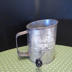 Pode incluir: Uma peneira de farinha de metal vintage com uma alça e um botão preto. A peneira tem formato cilíndrico com faixas horizontais e texto indicando as medidas. A alça é curva e a peneira é feita de metal prateado.