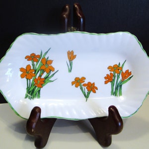 Pode incluir: Prato de servir retangular branco com borda ondulada e acabamento verde. O prato é decorado com ilustrações de flores laranja e caules verdes. O prato é exibido em um suporte de madeira marrom.