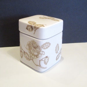 May include: A white, square ceramic container with a lid, decorated with a sepia-toned rose and leaf design. The container has a slightly rounded top and is set against a neutral background.