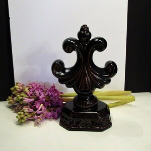 May include: A dark brown, decorative fleur-de-lis sculpture with a glossy finish. The base is octagonal and features an ornate design. The sculpture is paired with a bouquet of purple hyacinth flowers and long green stems.