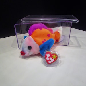 Puede incluir: Un juguete de peluche con forma de pez colorido con detalles en rosa, naranja, azul y morado. El pez tiene boca roja y ojos negros. Una etiqueta en forma de corazón con el texto "Ty" está adjunta. El juguete está dentro de una vitrina de plástico transparente.