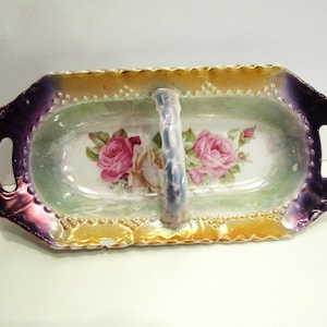 Pode incluir: Uma travessa de porcelana vintage com uma pega. A travessa tem formato retangular com borda ondulada e acabamento iridescente roxo e dourado. O interior apresenta um desenho floral com rosas cor-de-rosa.