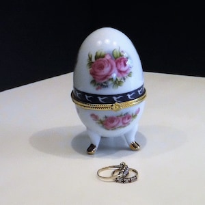 Puede incluir: Una caja de baratijas blanca de porcelana en forma de huevo con diseños florales de rosas rosas y detalles dorados. La caja tiene tres patas pequeñas y una tapa con bisagras. Dos anillos están delante de la caja.