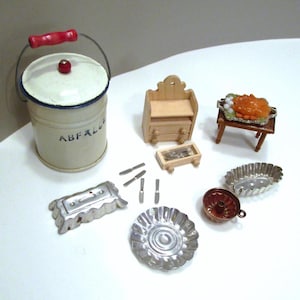 May include: A collection of miniature dollhouse kitchen items. Includes a cream-colored bucket with a red handle and the word "ABFALL", a small wooden cabinet, a table with a dish of food, and various metal molds and utensils.