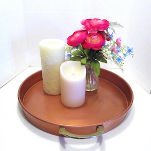 May include: A round copper-colored tray with brass handles holds two white candles and a small vase of pink and blue flowers. One candle is lit, and the other has a decorative pattern.