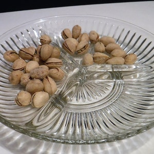 May include: A clear, divided glass serving dish filled with pistachios. The dish has three sections and a ribbed design. The pistachios are in their shells and have a light brown color.